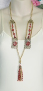 image shows a long statement tassel necklace, made from panels of filigree metal, studded with faux coral stones and tassels